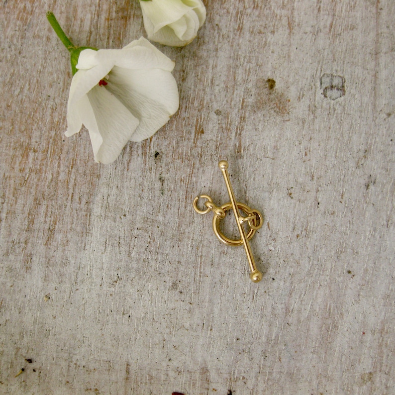 May include: Gold-toned metal toggle clasp with a round ring and a straight bar. The clasp is on a light wood surface.