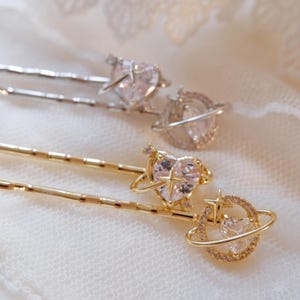 May include: Three hair pins with celestial designs. Two pins are silver-toned, and one is gold-toned. Each pin features a heart-shaped crystal and a ring of smaller crystals. The pins are set against a white, lacy background.