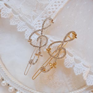 Op de afbeelding: Twee haarclips in de vorm van G-sleutels, versierd met strass en kleine vlinderaccenten. Eén clip is zilverkleurig, de andere goudkleurig. Ze liggen op een wit kanten oppervlak, met een parel- en strass-rand.