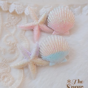 May include: Two starfish and two seashells with a glittery finish. One starfish is pink and white, while the other is purple and white. One seashell is pink and white, and the other is white and blue. The text The Snow Ball is in the corner.