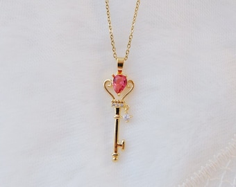 May include: A gold key necklace with a pink gemstone and clear crystal accents. The key has a crown-like top and a small star charm.