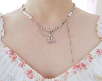 May include: A silver-toned necklace featuring a cherry pendant with pink accents. The necklace has a pearl strand and a delicate chain. The Snow Ball logo is visible in the bottom right corner. The necklace is worn over a floral patterned dress.