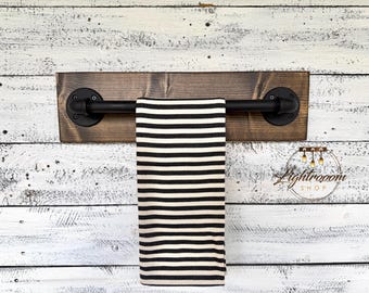 May include: A wall-mounted towel rack featuring a horizontal black metal pipe attached to a rectangular dark brown stained wooden board, with a black and white horizontally striped hand towel draped over the center of the pipe.