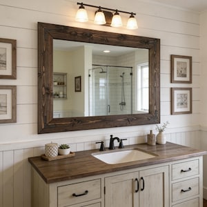 May include: A rustic bathroom vanity with a large, dark-framed mirror. The vanity has a light-colored cabinet with black hardware, a wooden countertop, and a white sink. Above the mirror is a bronze light fixture with three lights.