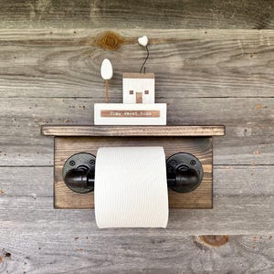 May include: A rustic wooden toilet paper holder with black pipe fittings. A small wooden house and a sign that reads "Home sweet home" are on a shelf above the roll of toilet paper. The background is weathered wood.