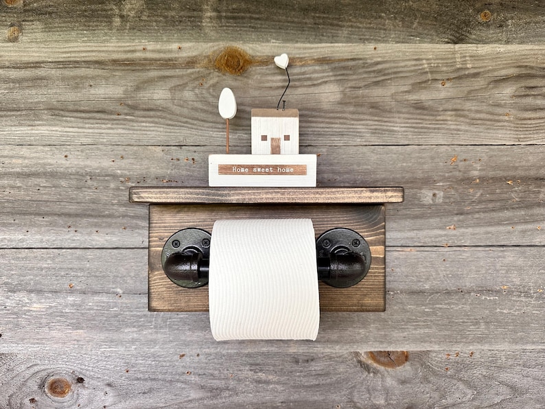 May include: A rustic wooden toilet paper holder with black pipe fittings. A small wooden house and a sign that reads "Home sweet home" are on a shelf above the roll of toilet paper. The background is weathered wood.