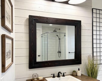 May include: Bathroom vanity with a dark wood cabinet, a white sink, and a large mirror with a dark wood frame. The mirror reflects a shower with a glass enclosure. Above the mirror are three black light fixtures.