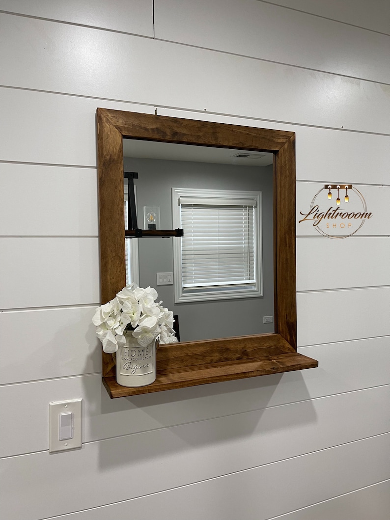 May include: A square, wooden-framed mirror with a shelf, mounted on a white shiplap wall. A white vase with the text "HOME where our story begins" holds white flowers. The mirror reflects a room with a window.