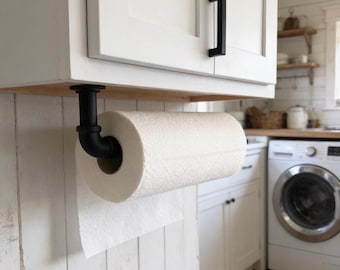 May include: A matte black industrial pipe paper towel holder mounted underneath a white kitchen cabinet. The holder features a sturdy L-shaped metal pipe design that securely supports a standard roll of white paper towels for easy access.