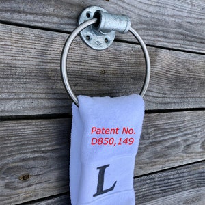 May include: A galvanized metal towel ring mounted to a wall, holding a white hand towel. The towel features the embroidered letter L and the red text Patent No. D850,149. The ring is constructed from industrial pipe fittings with a circular base.