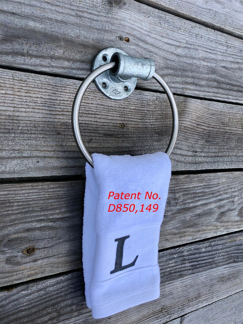 May include: A galvanized metal towel ring mounted to a wall, holding a white hand towel. The towel features the embroidered letter L and the red text Patent No. D850,149. The ring is constructed from industrial pipe fittings with a circular base.