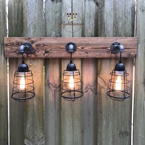 May include: Three industrial-style pendant lights with black metal cage shades and Edison bulbs are mounted on a weathered wooden plank. The lights are attached with metal pipe fittings. The background is a wooden fence.