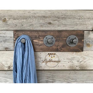 May include: A rustic wall-mounted coat rack made of dark stained wood, featuring three industrial-style metal hooks. A light blue scarf hangs from one hook. The rack is mounted on a weathered wooden wall.