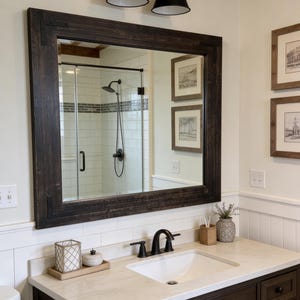 May include: A dark-stained wooden framed mirror hangs above a bathroom vanity. The vanity has a white countertop, a white sink, and dark brown cabinets. A black faucet and a pair of black light fixtures are also visible.