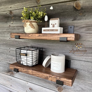 May include: Two dark brown wooden floating shelves mounted with black metal brackets. The top shelf holds a cream-colored Ball mason jar with greenery and a white house ornament labeled home sweet home. The bottom shelf holds a black wire basket.