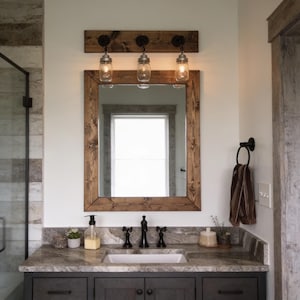 May include: A bathroom vanity set featuring a rectangular mirror with a dark stained wood frame, mounted below a matching three-light fixture with clear glass mason jar shades. The vanity includes a stone countertop and dark cabinets.