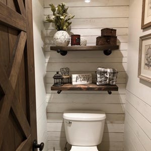 May include: A rustic bathroom scene with a white toilet and wooden shelves. The shelves hold decorative items like a vase with flowers, candles, and a sign that says "Pray". The walls are white with horizontal wood paneling, and a wooden door is visible on the left.