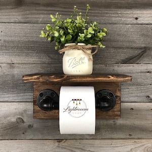May include: A rustic wooden toilet paper holder with a small shelf, featuring black pipe fittings. A white mason jar with the word "Ball" and greenery sits on the shelf. The background is weathered wood.
