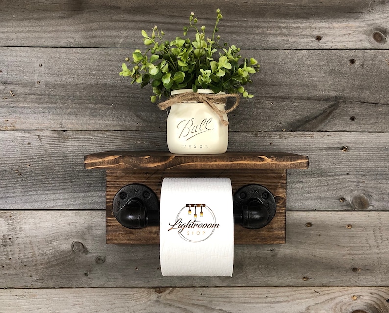 May include: A rustic wooden toilet paper holder with a small shelf, featuring black pipe fittings. A white mason jar with the word "Ball" and greenery sits on the shelf. The background is weathered wood.