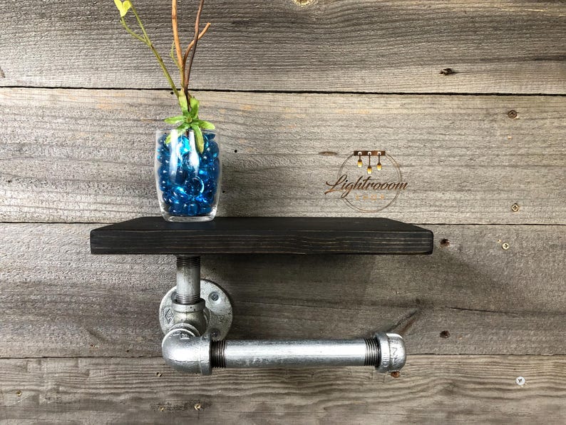 Puede incluir: Un estante de pared con una tabla de madera te&ntilde;ida de oscuro, sostenida por un soporte de tuber&iacute;a de metal industrial de tono plateado. Un florero de vidrio transparente con cuentas azules y una peque&ntilde;a planta verde descansa sobre &eacute;l.
