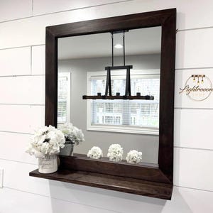 May include: A rectangular dark brown framed mirror with a shelf, mounted on a white shiplap wall. The mirror reflects a room with a window and a black light fixture. White flowers in a white pot and three smaller arrangements sit on the shelf.