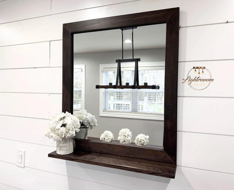 May include: A rectangular dark brown framed mirror with a shelf, mounted on a white shiplap wall. The mirror reflects a room with a window and a black light fixture. White flowers in a white pot and three smaller arrangements sit on the shelf.