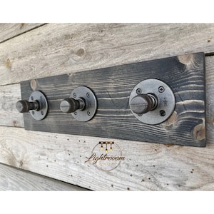 May include: A wall-mounted coat rack featuring a rectangular, dark-stained wooden board with visible grain. Three industrial-style metal pipe hooks are attached to the board, each secured with a circular flange and four screws. The logo Lightrooom SHOP is visible.