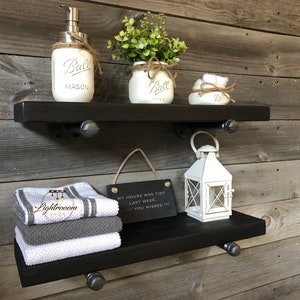 May include: Two black floating shelves with industrial pipe brackets are mounted on a wooden wall. The top shelf holds a soap dispenser, a small plant in a mason jar, and a decorative item. The bottom shelf displays folded towels, a white lantern, and a small sign that reads "MY HOUSE WAS TIDY LAST WEEK. SORRY YOU MISSED IT!"