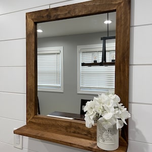 May include: A square, wooden-framed mirror with a shelf, mounted on a white wall. The mirror has a dark brown finish. A white vase with white flowers and the text "HOME where our story begins" sits on the shelf.