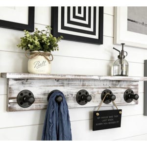 May include: A rustic, white-washed wooden shelf with four black metal hooks. A blue scarf hangs from one hook. A small sign reads "My house was tidy last week, sorry you missed it!" A mason jar with greenery sits on the shelf.