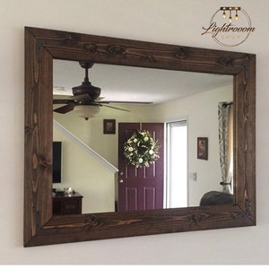 May include: A rectangular mirror with a dark brown wooden frame. The frame has visible wood grain and is constructed with corner brackets. The mirror reflects a room with a ceiling fan, door, and a white floral wreath.