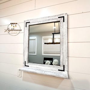 May include: Square mirror with a distressed white wooden frame and black corner accents. The mirror is mounted on a white wall with horizontal paneling. The reflection shows a room with a window and a black chair. The mirror has a shelf.