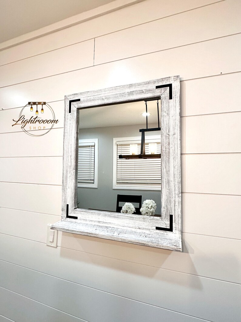 May include: Square mirror with a distressed white wooden frame and black corner accents. The mirror is mounted on a white wall with horizontal paneling. The reflection shows a room with a window and a black chair. The mirror has a shelf.