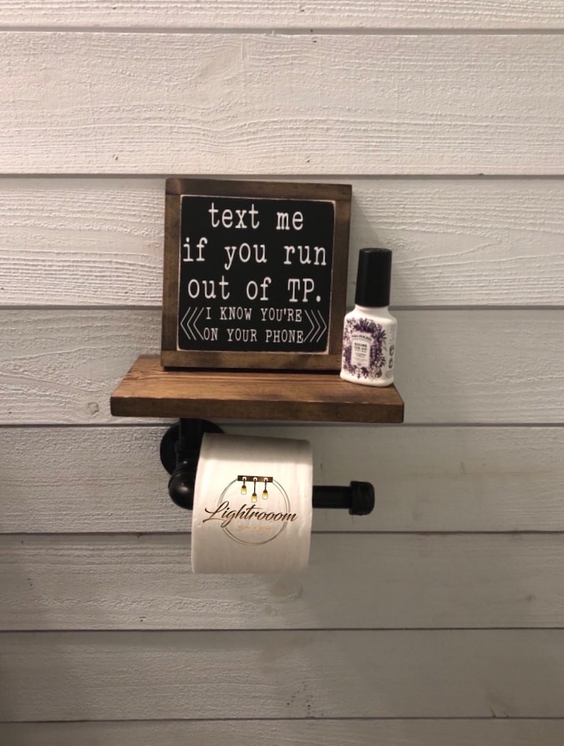 Puede incluir: Un soporte para papel higi&eacute;nico montado en la pared hecho de tuber&iacute;a industrial negra con un estante de madera oscura. Un letrero de madera cuadrado con texto blanco dice: text me if you run out of TP. I KNOW YOU'RE ON YOUR PHONE. Un rollo de papel y un frasco.