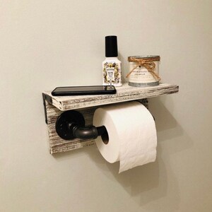 May include: A rustic, wall-mounted toilet paper holder with a shelf. The shelf is made of distressed white wood and features a black pipe for the toilet paper roll. A black smartphone, a bottle, and a candle sit on the shelf.
