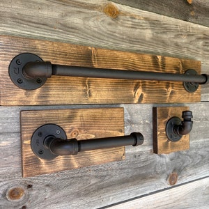 Puede incluir: Un juego de tres accesorios de ba&ntilde;o con tuber&iacute;as de metal negro mate montadas sobre tablas de madera te&ntilde;ida rectangulares. El conjunto incluye una barra larga para toallas, un portarrollos de papel higi&eacute;nico y un gancho individual.