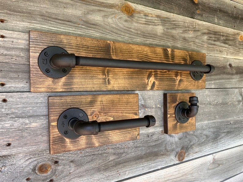 Puede incluir: Un juego de tres accesorios de ba&ntilde;o con tuber&iacute;as de metal negro mate montadas sobre tablas de madera te&ntilde;ida rectangulares. El conjunto incluye una barra larga para toallas, un portarrollos de papel higi&eacute;nico y un gancho individual.