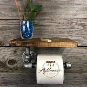 Puede incluir: Un soporte de papel higi&eacute;nico montado en la pared hecho de tuber&iacute;as de metal gris industrial con un estante de madera marr&oacute;n oscuro en la parte superior. Un rollo de papel higi&eacute;nico blanco con el texto Lightrooom SHOP impreso cuelga de la tuber&iacute;a. Un jarr&oacute;n de vidrio con piedras azules y una planta verde se asienta sobre el estante.