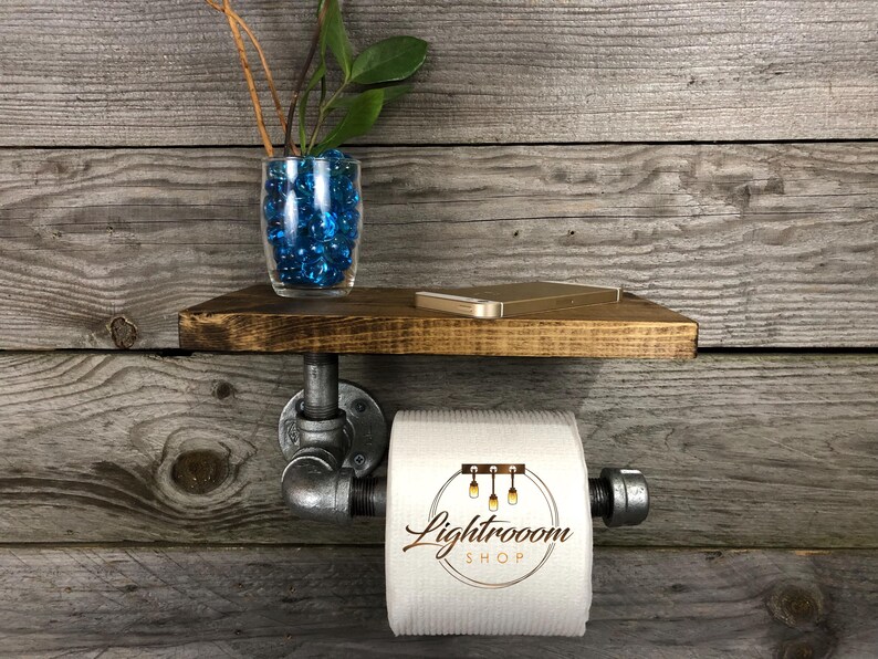 Puede incluir: Un soporte de papel higi&eacute;nico montado en la pared hecho de tuber&iacute;as de metal gris industrial con un estante de madera marr&oacute;n oscuro en la parte superior. Un rollo de papel higi&eacute;nico blanco con el texto Lightrooom SHOP impreso cuelga de la tuber&iacute;a. Un jarr&oacute;n de vidrio con piedras azules y una planta verde se asienta sobre el estante.