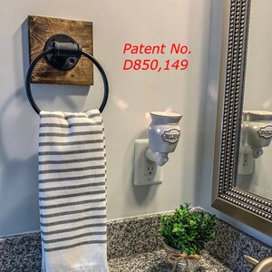 May include: A wall-mounted towel ring featuring a black metal circular holder attached to a square dark wood base. A white and gray striped hand towel hangs from the ring. The text Patent No. D850,149 is visible on the wall near the towel ring.