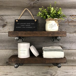 May include: Two-tier rustic wooden shelves with black pipe brackets. The top shelf holds a sign that reads "MY HOUSE WAS TIDY LAST WEEK. SORRY YOU MISSED IT!" and a Ball jar with greenery. The bottom shelf has a candle and a folded towel.