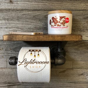 Puede incluir: Un soporte para papel higi&eacute;nico de pared hecho de tuber&iacute;a industrial negra con un estante de madera oscura. Un rollo de papel higi&eacute;nico blanco con el logotipo de Lightrooom Shop est&aacute; en el soporte. Una vela blanca etiquetada Gingerbread Cookies est&aacute; en el estante.