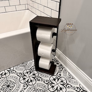 May include: A dark brown wooden toilet paper holder with three rolls of white toilet paper. The holder has a rectangular design and black metal rods to hold the rolls. The holder is in a bathroom with black and white patterned floor tiles.