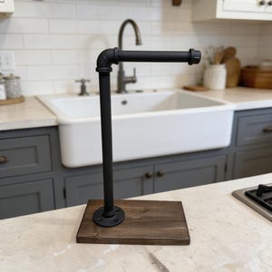 May include: A paper towel holder featuring a matte black metal pipe assembly attached to a rectangular dark brown wooden base. The vertical pipe connects to a horizontal arm via an elbow fitting, creating an L-shaped structure for holding rolls.