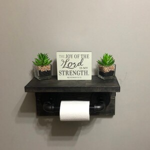 May include: A rustic, dark-stained wooden shelf with a built-in toilet paper holder. The shelf features a small sign with the text "THE JOY OF THE LORD IS MY STRENGTH." Two small potted succulents sit on the shelf.