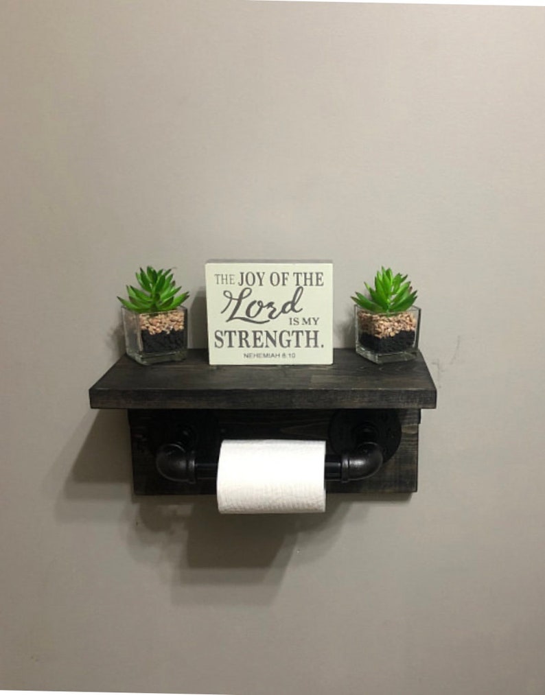 May include: A rustic, dark-stained wooden shelf with a built-in toilet paper holder. The shelf features a small sign with the text "THE JOY OF THE LORD IS MY STRENGTH." Two small potted succulents sit on the shelf.