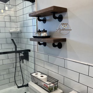May include: Two rustic wooden shelves with black pipe brackets mounted on a white tiled wall. The shelves are stained a dark brown and appear to be in a bathroom setting. The shelves are holding jars and a black tray.