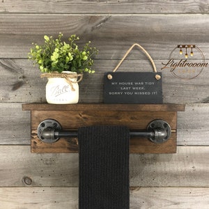May include: A dark wood wall shelf featuring a black metal pipe towel bar, a white Ball mason jar with green foliage, and a rectangular black slate sign with the text: MY HOUSE WAS TIDY LAST WEEK. SORRY YOU MISSED IT! A black towel hangs from the bar.