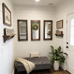 May include: A gray tufted bench with a cream throw blanket and a pillow reading farmhouse ish, positioned below three rectangular dark wood mirrors, two wooden wall shelves with hooks, and framed wall art on white shiplap walls.