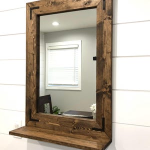May include: A rectangular mirror with a dark brown wooden frame and a shelf. The frame has black metal corner brackets. The mirror is mounted on a white wall with horizontal panelling.